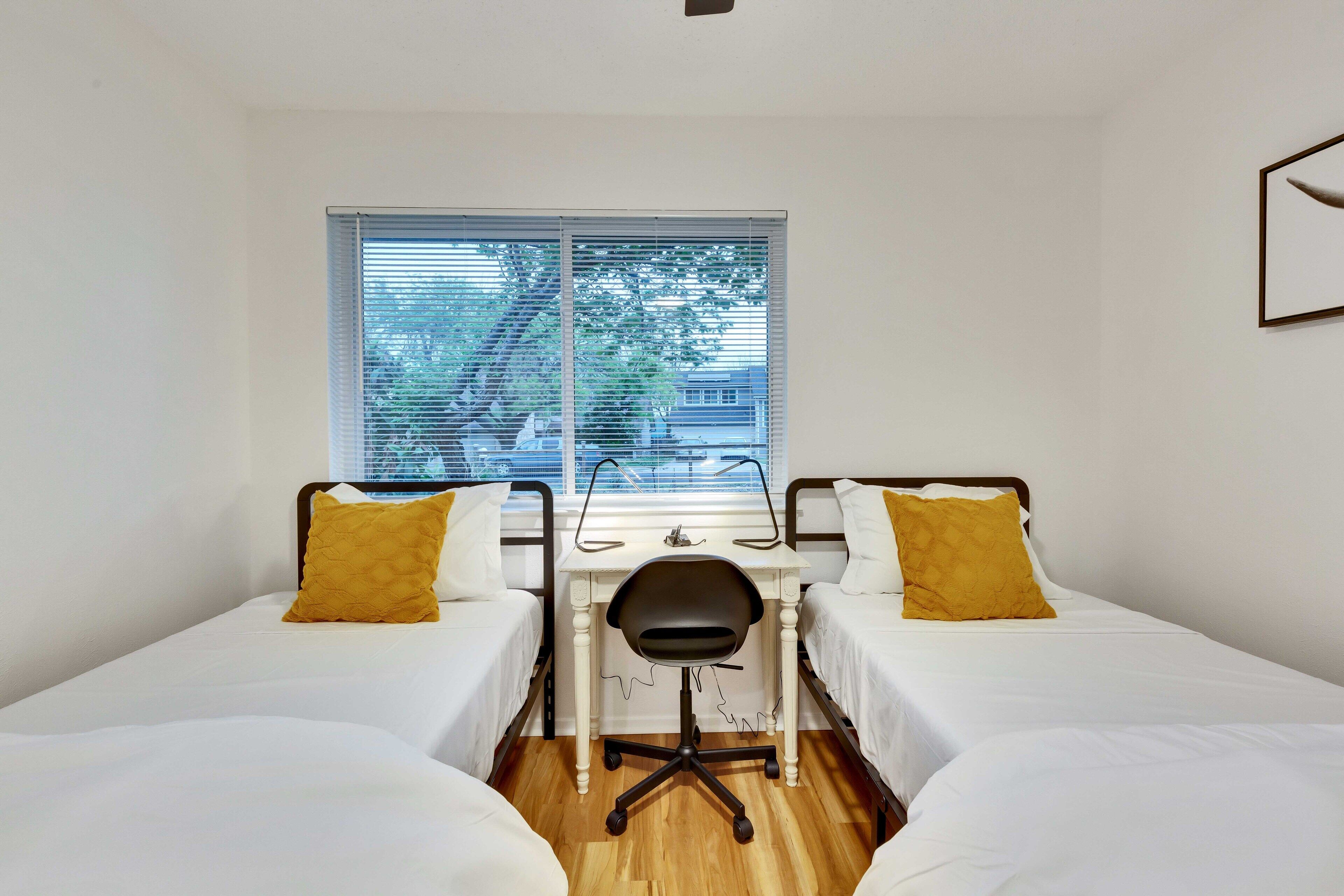 A room with two beds, a desk, and a chair. There is a window with blinds, and a tree is visible outside.