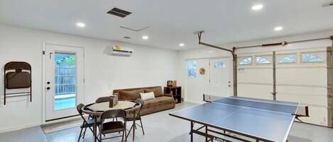 A room with a ping pong table, a sofa, a dining table with chairs, and a door leading outside.