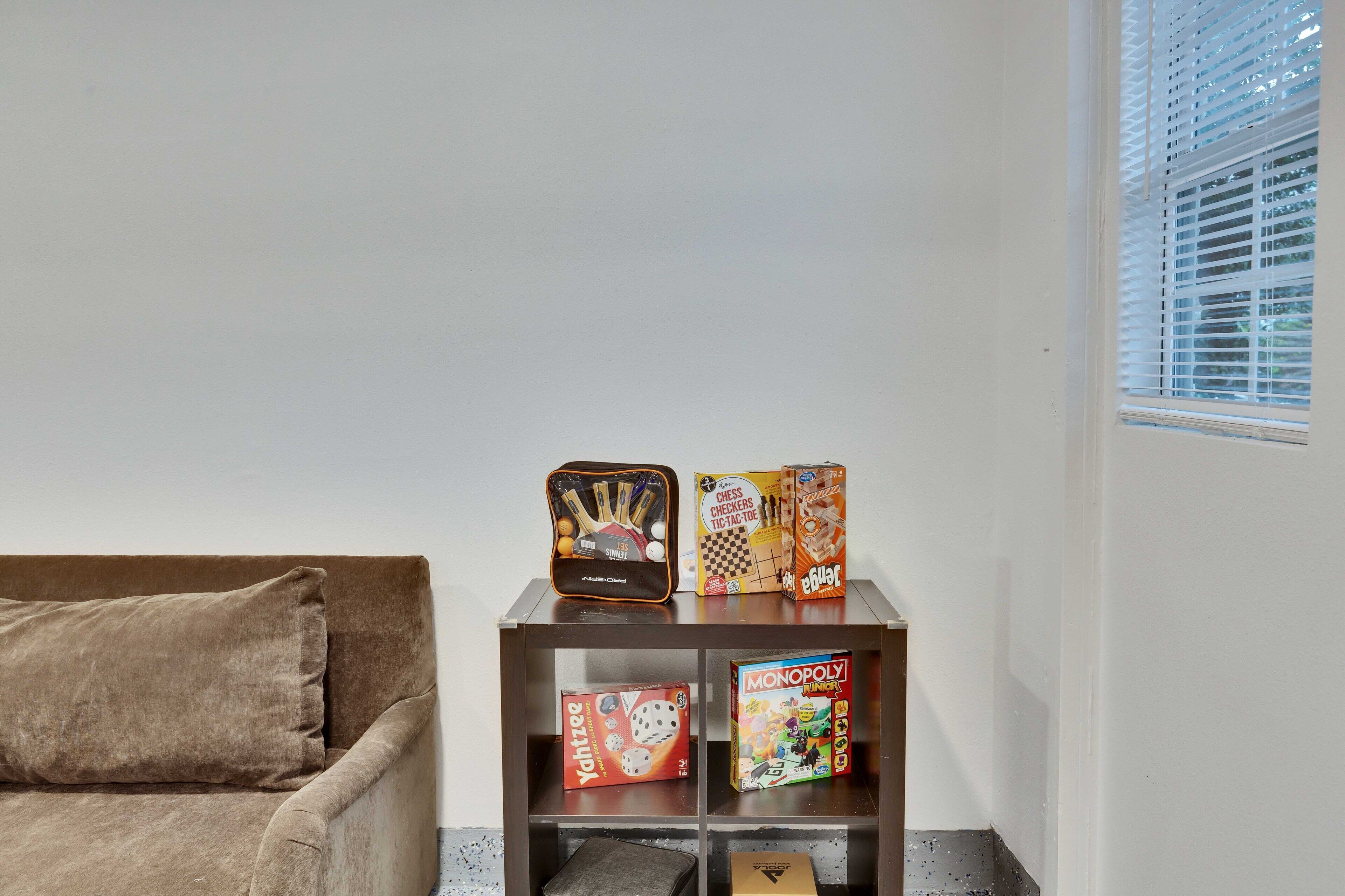 A brown sofa with a cushion, a shelf with board games, and a window with blinds.