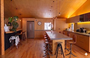 A modern kitchen with wooden cabinets, a central island with stools, a refrigerator, and a clock on the wall.