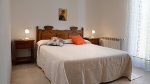 A neatly made bed with a floral pillow and an orange accent pillow, flanked by two bedside tables with lamps, a radiator, and a window with sheer curtains.