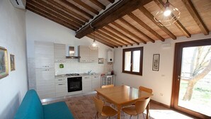 Une cuisine avec des poutres en bois, des armoires blanches, une table à manger avec des chaises et une vue sur l’extérieur par une fenêtre.