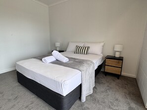 Un lit bien fait, avec du linge de lit blanc, un plaid gris et deux serviettes enroulées. Il y a une table de chevet avec une lampe et un petit tiroir.