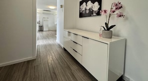 Un couloir avec une console blanche, une orchidée en pot et un tableau représentant une orchidée en noir et blanc accroché au mur.