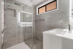 Een moderne badkamer met een glazen douchecabine, een witte wastafel en een spiegel met een raam erboven.