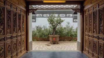 An indoor corridor with intricate wooden carvings on the walls and ceiling, leading to a courtyard with a potted tree.