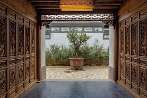 An indoor corridor with intricate wooden carvings on the walls and ceiling, leading to a courtyard with a potted tree.