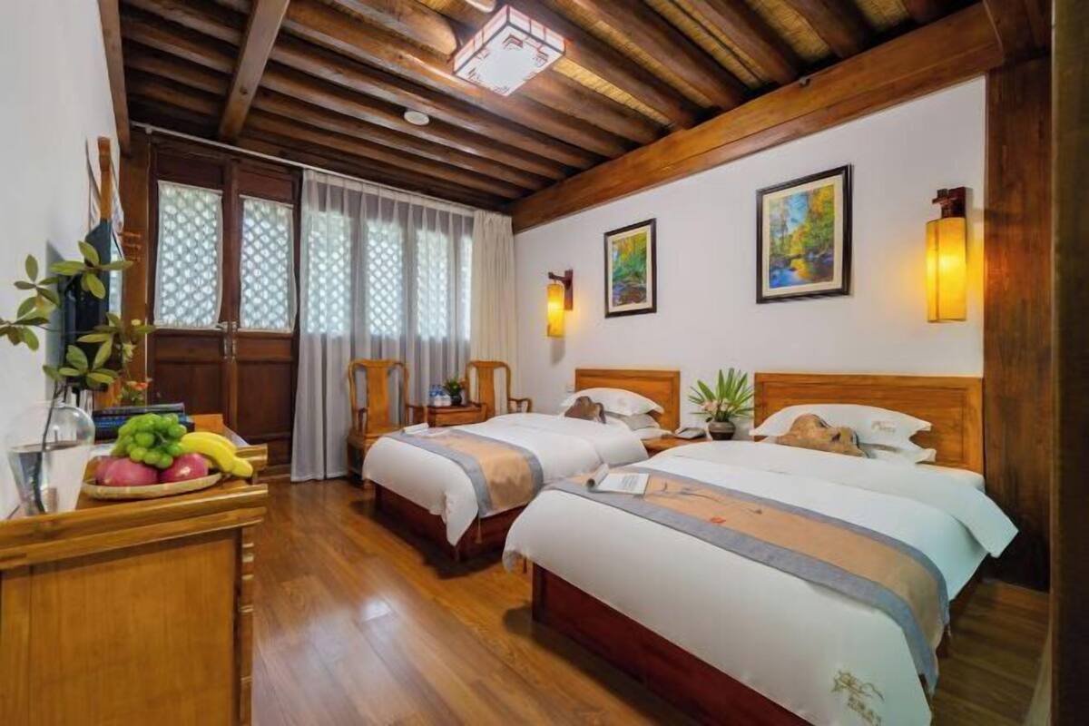 A hotel room with two beds, wooden flooring, a desk with fruit, a window with curtains, and wall-mounted lights.