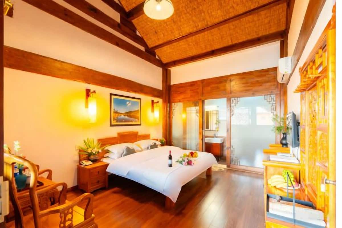 A hotel room with a wooden ceiling, a bed with a white bedspread, a nightstand with a lamp, a wooden chair, and a sliding glass door leading to a balcony.