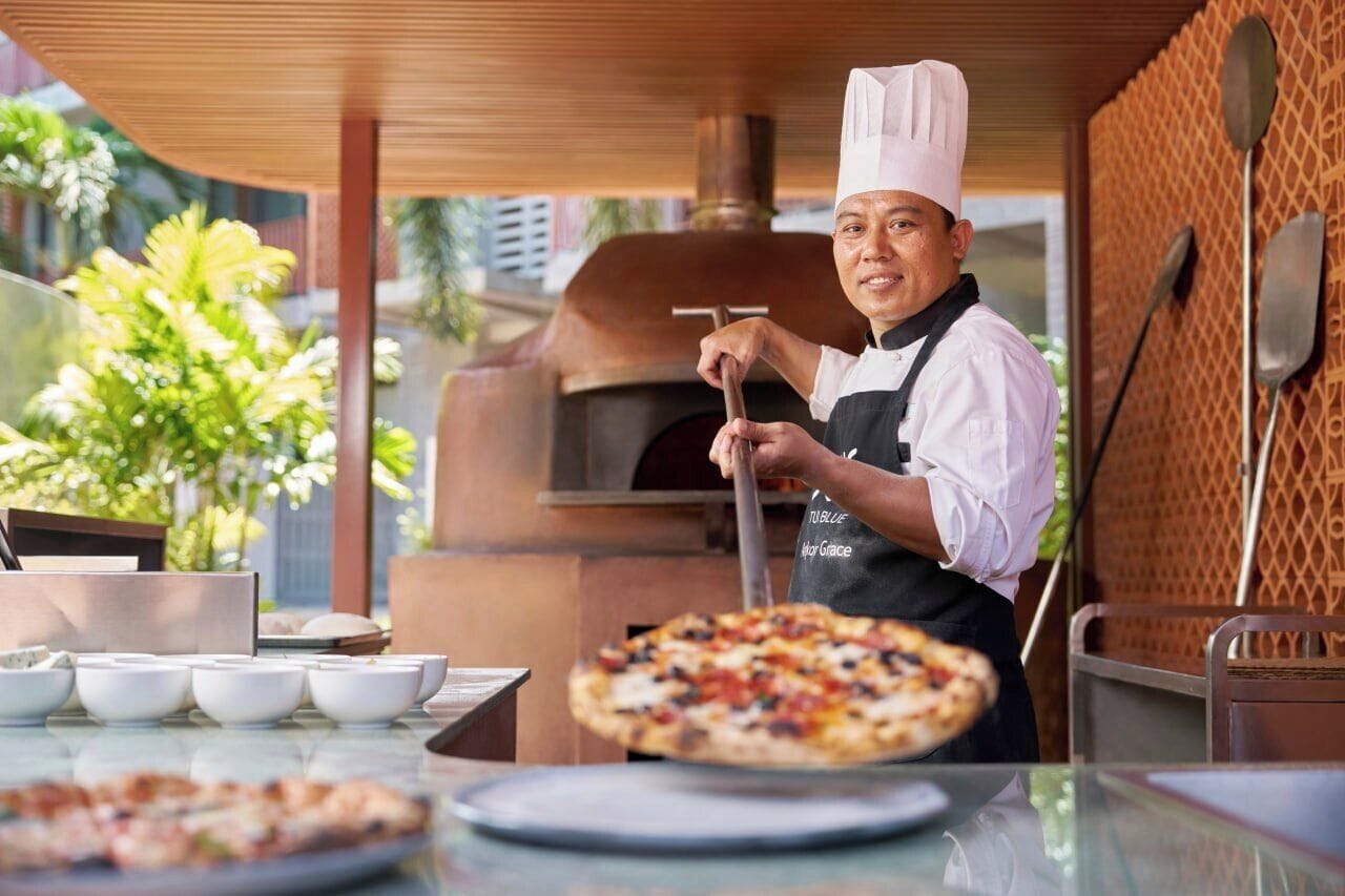 A chef in a white chef's hat and apron, preparing a pizza in an outdoor brick oven.