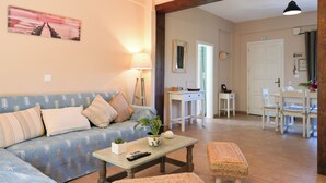 Ein Wohnzimmer mit Sofa, Couchtisch und Hocker, ein Essbereich mit Tisch und Stühlen sowie eine durchgehende Tür erkennbare Küche.