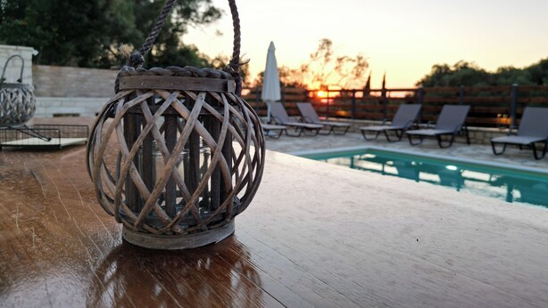 Ein Korbstuhl-Laterne steht auf einem Holztisch in der Nähe eines Pools mit Liegestühlen, im Hintergrund ein Sonnenuntergang.