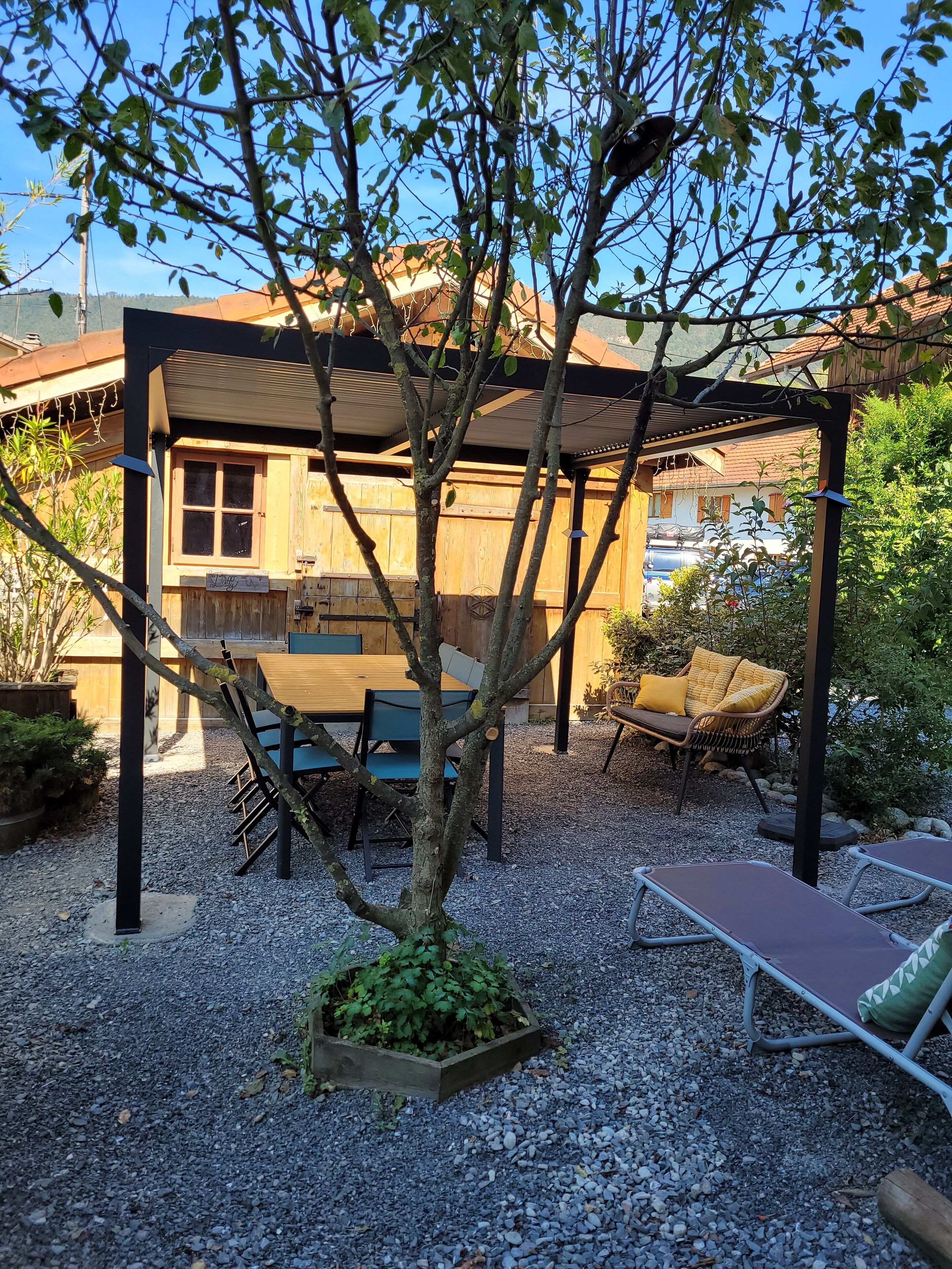 Een overdekt terras met een tafel, stoelen, een kleine boom en een gebouw met een houten gevel op de achtergrond.