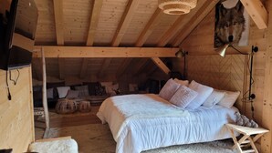 Une chambre mansardée chaleureuse avec un plafond en bois, un lit recouvert de linge de maison blanc, une petite table en bois et une lampe fixée au mur.