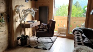 L’intérieur d’un chalet en bois chaleureux et accueillant, avec un canapé, un fauteuil, une petite table agrémentée d’une plante, et une vue sur la verdure à l’extérieur.