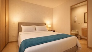 A hotel room with a bed, a nightstand, a lamp, a bathroom with a sink and mirror, and a door leading to another room.