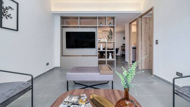 A modern living room with a large flat-screen TV, a grey sofa, a coffee table with magazines, and a decorative vase.