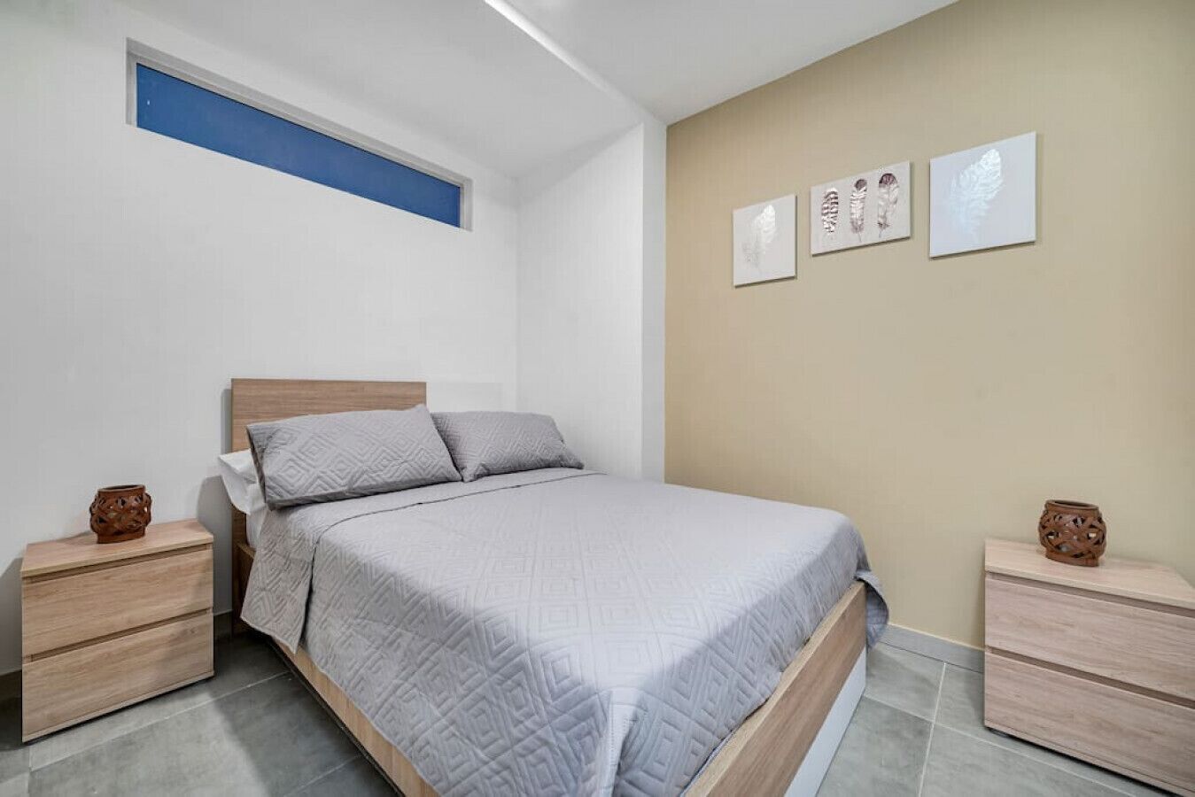 A bedroom with a bed, two nightstands, a window with a blue frame, and three wall-mounted artworks.