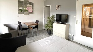 A modern living room with a sofa, a dining table with chairs, a TV on a cabinet, and a door leading to another room.