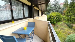A balcony with a table and chairs, a yellow glass, and a view of a garden with trees and shrubs.