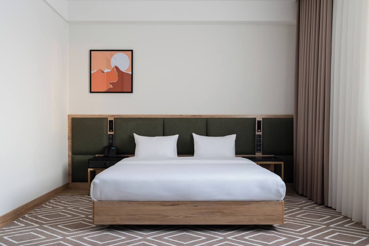 A hotel room with a large bed, a headboard, a bedside table with a phone, and a wall-mounted artwork.