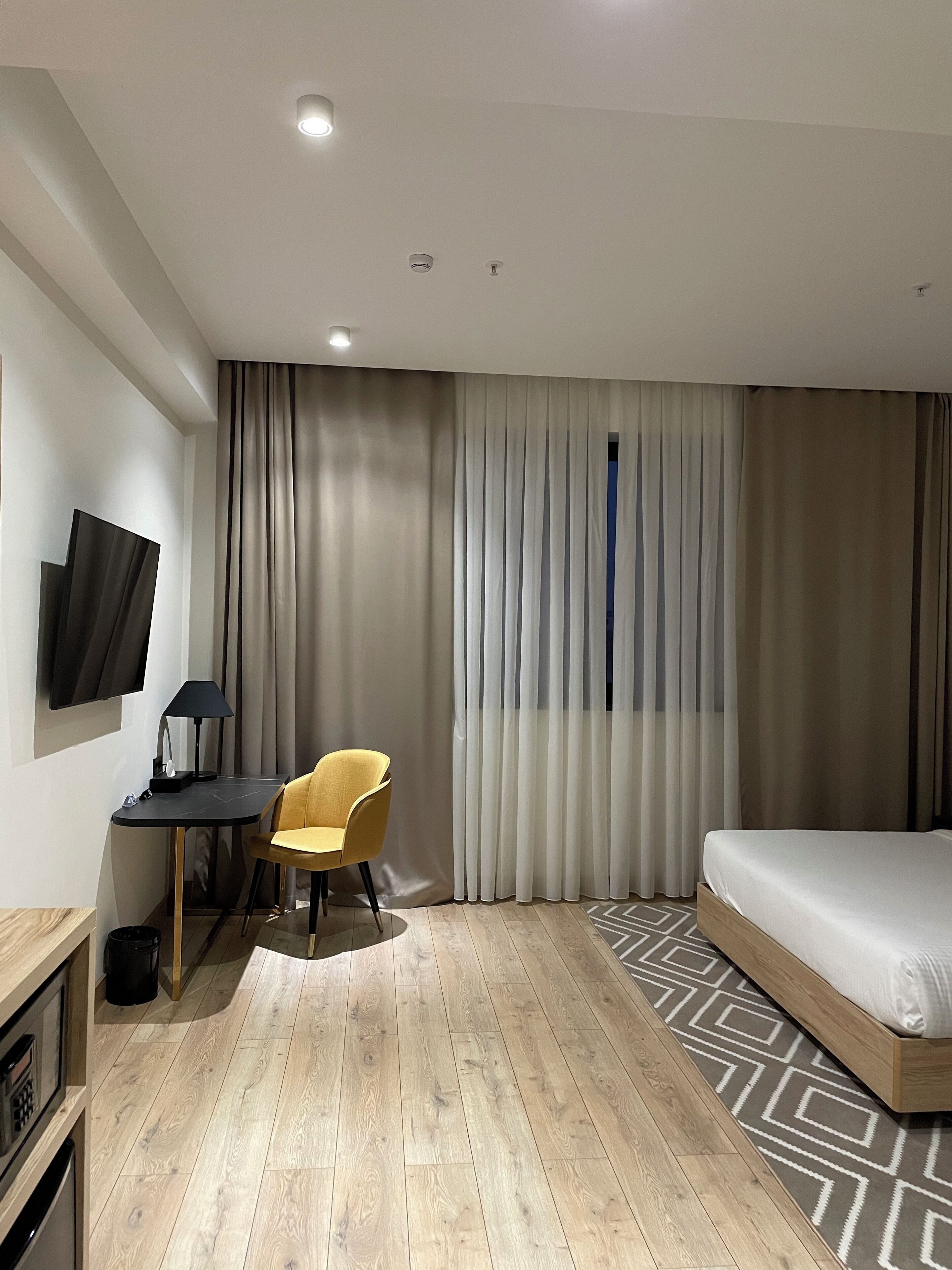 A modern hotel room with a wooden floor, a bed, a desk with a chair, a TV mounted on the wall, and a window with curtains.