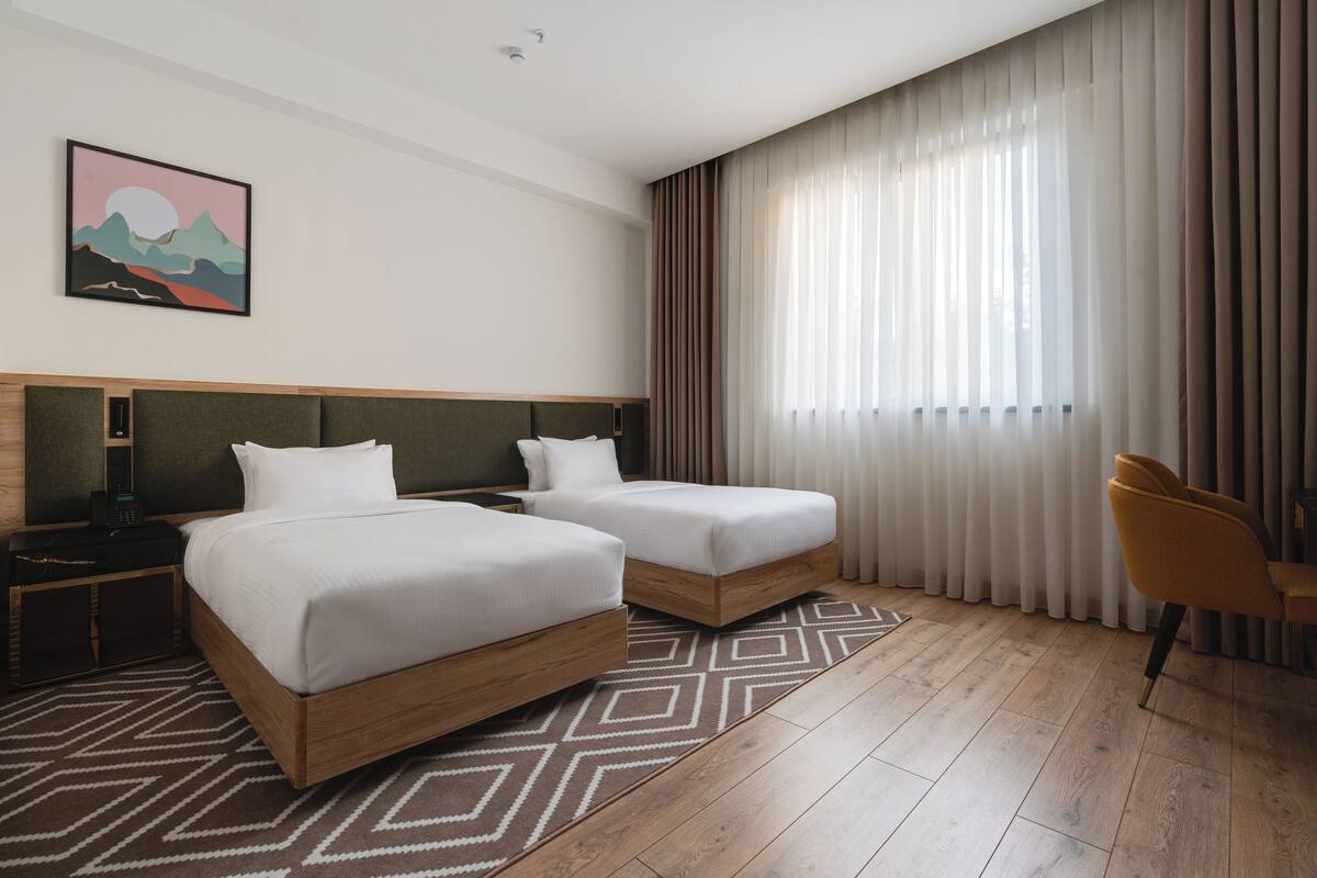 A hotel room with two beds, a wooden headboard, a nightstand, a chair, and a large window with curtains.