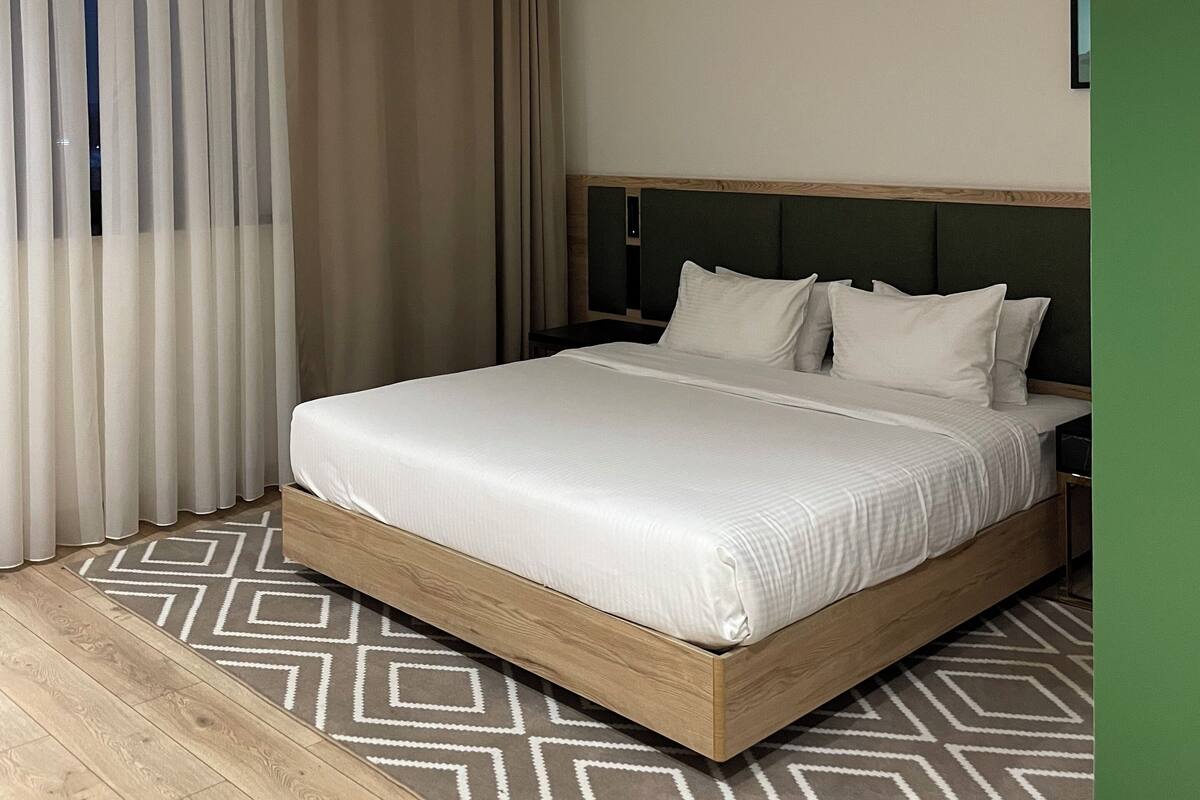 A modern hotel room with a large bed, wooden headboard, and a geometric patterned rug.