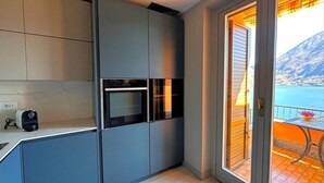 Una cocina moderna con armarios grises, horno empotrado y una pequeña encimera. Hay una puerta corrediza de vidrio que da a un balcón con vistas al mar y a las montañas.