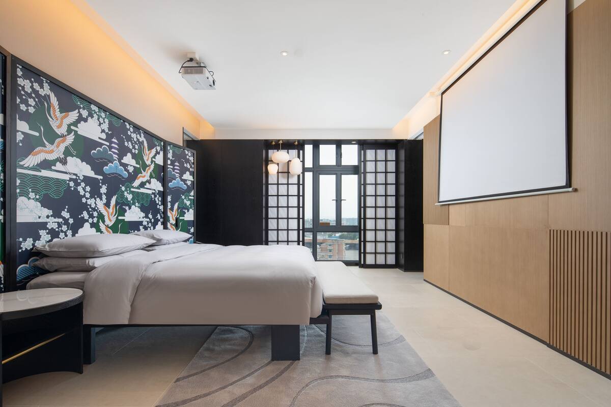 Ein modernes Hotelzimmer mit einem großen Bett, einem kleinen Nachttisch, einer Bank, einem großen Fenster mit Blick und einer projizierten Leinwand an der Wand.