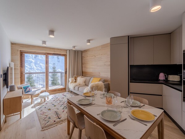 A modern, well-lit dining area with a table set for four, a sofa with a view of the mountains, and a kitchen area with a TV and appliances.