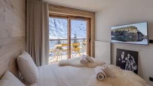 A hotel room with a bed, a large window with a view of snow-covered mountains, a balcony with yellow chairs, and a television on the wall. - Modern 2-room apartment, near slopes, Courchevel (Courchevel 1650)