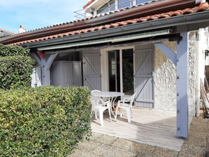 A covered outdoor seating area with a table and chairs, a wooden deck, and a white building with a tiled roof.