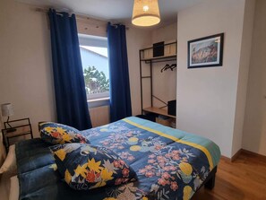 A bedroom with a bed, a window with curtains, a bedside table, a lamp, a shelf with clothes, and a framed picture on the wall.