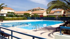 Une piscine avec un bordure bleue, entourée d’une petite clôture. On aperçoit des palmiers et un bâtiment avec un toit en tuiles à l’arrière-plan.