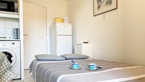 Une chambre à coucher avec un lit, des oreillers, une table de chevet avec une tasse et une soucoupe, une petite armoire blanche et un lave-linge.
