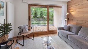 A modern living room with a grey sofa, a small wooden table, and a chair. There is a large sliding glass door leading to an outdoor area with a wooden deck and a view of trees.