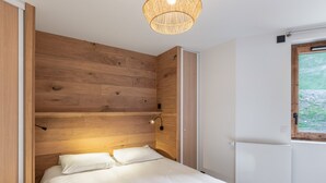 A bedroom with a wooden headboard, a bed with white bedding, a hanging light fixture, and a window with a view of greenery.