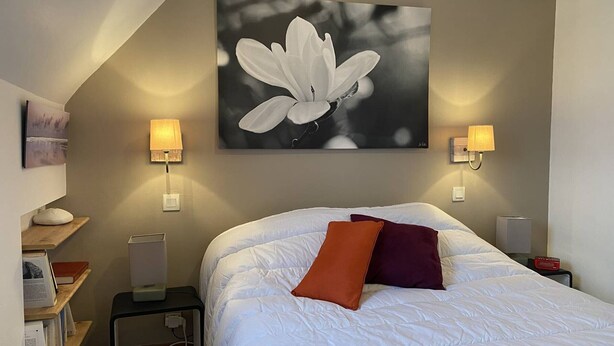 Un dormitorio con una cama grande, dos lámparas de noche, un cuadro colgado en la pared de una flor, una mesita de noche con una lámpara, y una estantería con libros.