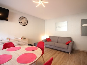 Ein modernes Wohnzimmer mit grauem Sofa, roten Sesseln, einem Holzesstisch mit roten Tischsets, einer Wanduhr, einem Flachbildfernseher und einem gerahmten Kunstwerk.