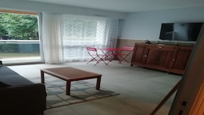 Un salon comprenant un canapé, une table en bois, un téléviseur et un coin cuisine avec des placards et une fenêtre.