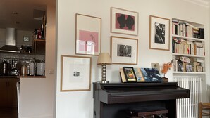 Un salon avec un piano, des œuvres d’art encadrées accrochées au mur, une bibliothèque et un radiateur.
