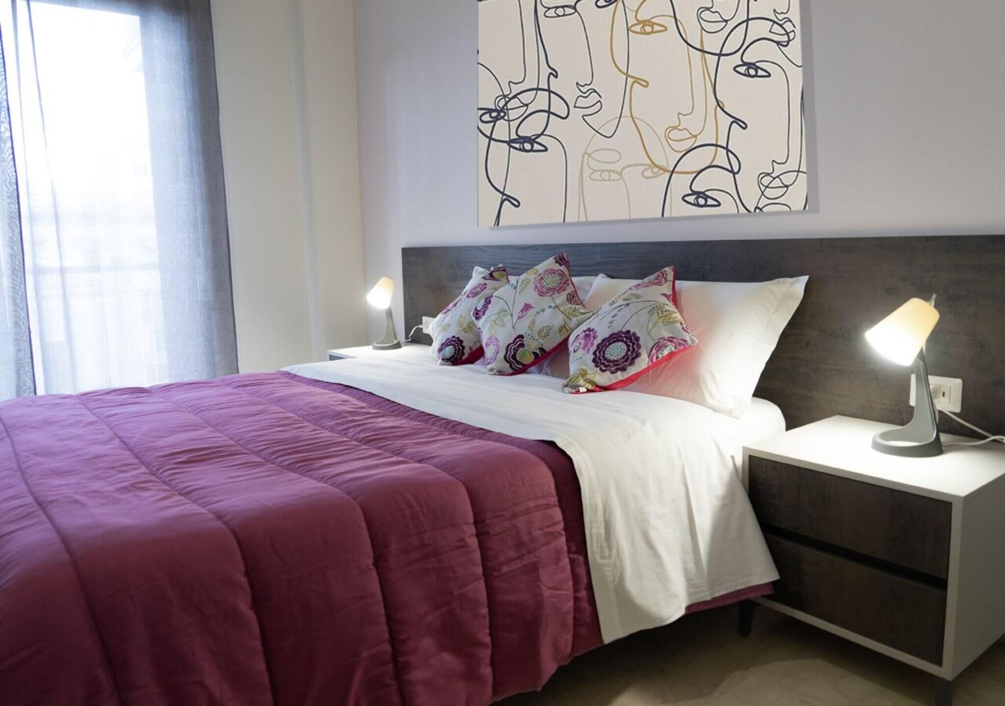 A neatly made bed with a red bedspread, a white pillow, and a decorative pillow with a floral pattern. There is a bedside table with a lamp and a wall art piece above the bed.
