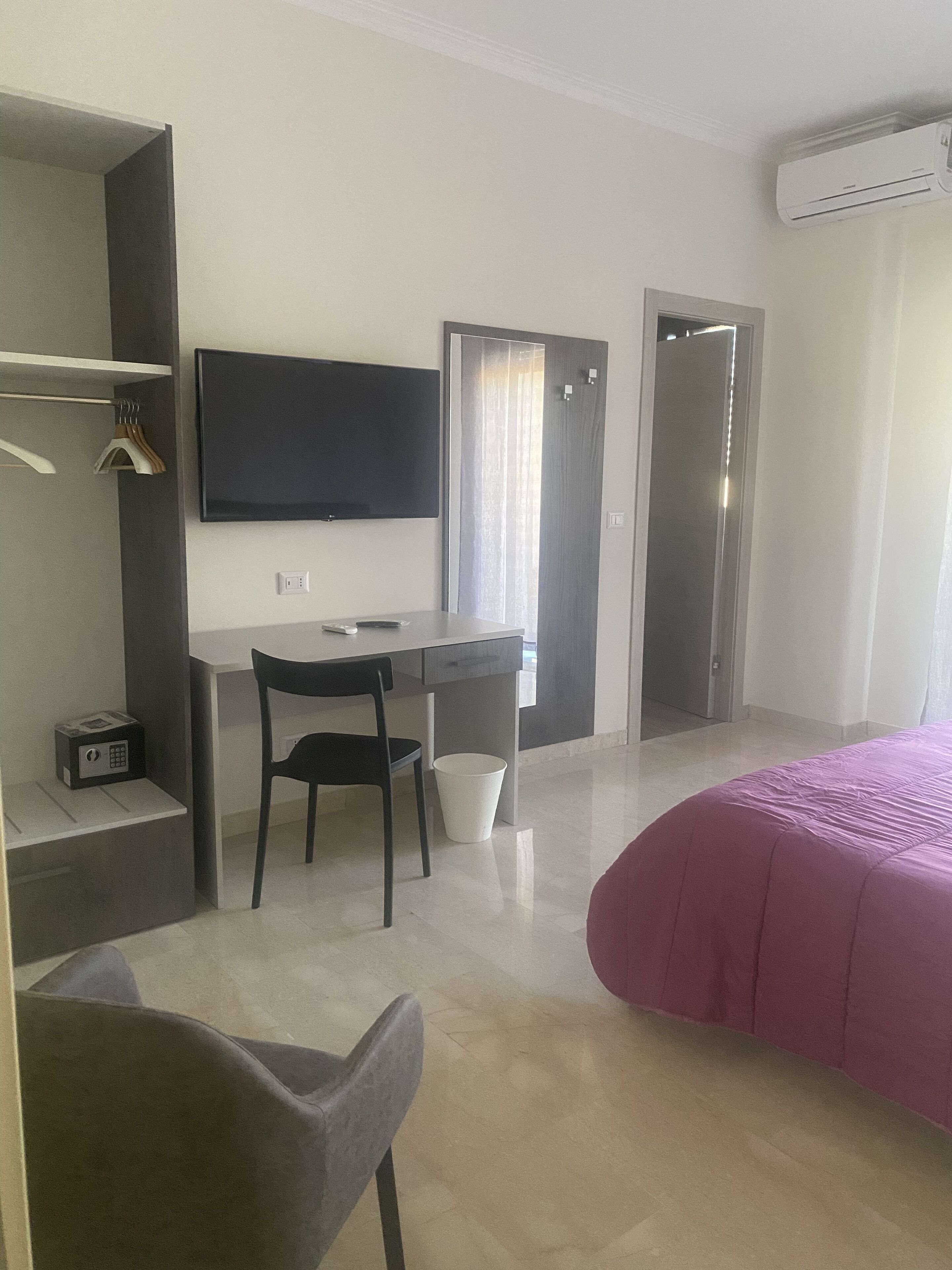 Una habitación de hotel moderna con escritorio, silla y cama con cubierta rosa. Hay un televisor montado en la pared, una puerta con espejo y un equipo de aire acondicionado en el techo.