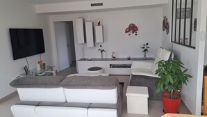 Ein modernes Wohnzimmer mit einem weißen Sofa, einem Couchtisch und einem an der Wand befestigten Fernseher. Ein kleiner Tisch mit Dekoration und einer Topfpflanze steht ebenfalls dort.