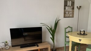 A living room with a TV on a wooden stand, a potted plant, a dining table with chairs, and a wall-mounted sign. - Charming, quiet duplex cottage with terrace/garden (Ardon)