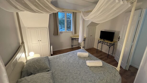 A bedroom with a canopy bed, a television on a side table, and a window with curtains. - Charming, quiet duplex cottage with terrace/garden (Ardon)