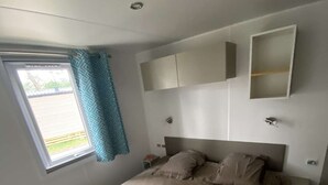 A bedroom with a bed, a window with curtains, a bedside table, and a wall-mounted cabinet. - Mobile home camping 5 stars (Vendays-Montalivet)