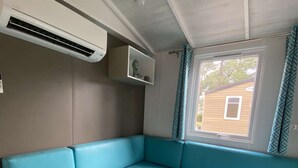 A room with a blue sofa, an air conditioning unit, a window with curtains, and a view of a building outside. - Mobile home camping 5 stars (Vendays-Montalivet)