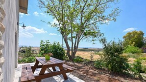 A wooden picnic table with a green bowl and pink flowers, set on a patio with a view of a tree and open fields. - YalaRent Valley view complex In Merhavia with Private heated Pool (Emek Izrael)
